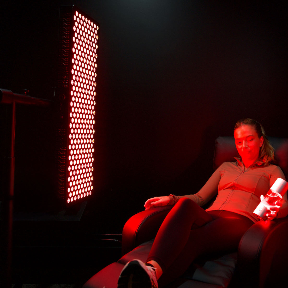 HUVE LUMA Restore Max in Black mounted to the HUVE Premium Floor Stand with fit woman relaxing on a chair with the HUVE Perform hydrogen water bottle